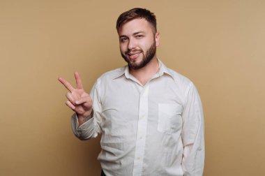 Bir adam sade bir arka planın önünde duruyor, gülümsüyor ve sağ eliyle barış işareti yapıyor. Beyaz bir gömlek giyiyor ve bu sıradan anda rahat görünüyor..