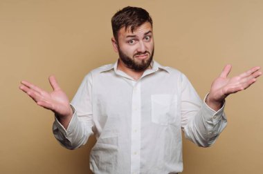 Bir adam yüzünde sorgulayıcı bir ifadeyle ellerini kaldırır. Sakalı var ve beyaz bir gömlek giyiyor. Arka plan sağlam bir renk. Sahne rahat ve rahat görünüyor..