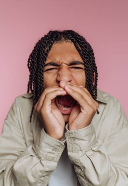 Genç bir adam ellerini ağzına koyarken bağırıyor. Kıvırcık saçları var ve açık renkli bir ceket giyiyor. Arka plan parlak pembe bir renktir..