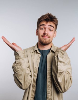 Genç bir adam arka planı hafif bir stüdyoda duruyor. Açık renk saçları ve sakalı var. Kafası karışmış bir ifadeyle ellerini kaldırır, sanki soru sorar gibi..