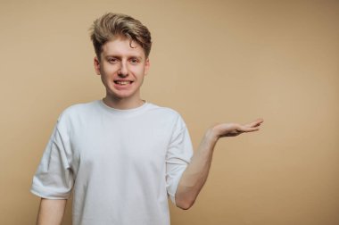 Genç bir adam elini dışarı uzatırken gülümsüyor. Düz beyaz bir gömlek giyiyor ve açık kahverengi bir duvara dayanıyor. Dekor bir stüdyo gibi görünüyor..