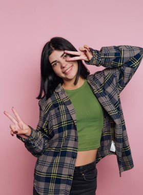 Genç bir kadın pembe bir duvarın önünde duruyor. Gülümsüyor ve yeşil bluz ve flanel gömlek giyerken eliyle barış işareti gösteriyor..