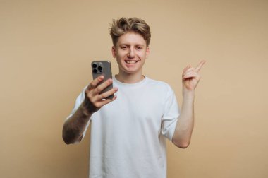 Genç bir adam içeride duruyor, bir elinde akıllı bir telefon, diğerini işaret ediyor. Kameraya bakarken gülümsüyor. Arkaplan sade ve açık renkli..