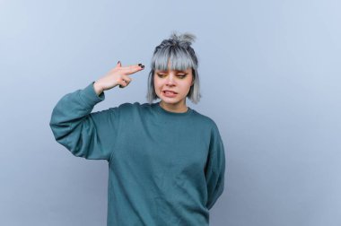 Genç bir kadın, bir eli havada bir silah hareketini taklit ederek ayakta duruyor. Kısa, açık renkli saçları var ve sade bir kazak giyiyor. Arka plan gri..