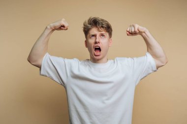 Bej bir geçmişi olan bir adam stüdyoda duruyor. Kollarını kaldırıyor ve kaslarını esnetiyor. Sanki bağırıyormuş gibi ağzı açık. Sahne coşkulu bir ifadeyi yakalıyor..