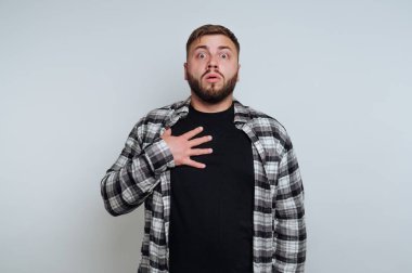 Bir adam stüdyoda duruyor ve şaşırmış bir ifade sergiliyor. O, kameraya doğrudan bakarken elini göğsüne koydu. Arkaplan sade ve hafif.