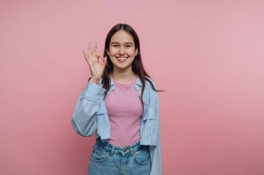 Bir kadın pembe arka planda duruyor. Pembe bir bluzun üzerine açık mavi bir gömlek giyiyor ve uzun saçları var. Gülümsüyor ve sağ eliyle 