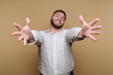 Kollarını açıp gülümseyen bir adam. Beyaz bir gömlek giyiyor ve hareketli bir andan hoşlanıyor gibi görünüyor. Arka plan basit bir bronzlaşmadır, bu da kapalı bir ortam olduğunu gösterir..