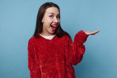 Bir kadın kırmızı bir kazak giyerken büyük bir gülümseme gösterir. Sanki ona katılması için birini davet ediyormuş gibi avucunu kaldırıyor. Ayarın düz mavi bir arkaplanı var.