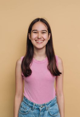 Bir kız sarı bir duvarın önünde poz veriyor. Pembe gömlek ve kot pantolon giyiyor. Onun saçı uzun ve düz. Kapalı bir ortamda, hafif ışıklandırmayla dururken gülümsüyor..