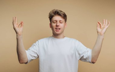 Genç bir birey, gözleri kapalı ve elleri sessiz bir yerde durarak farkındalık egzersizleri yapıyor. Arka plan sade ve tarafsız. Kişiye odaklanmamızı sağlıyor..