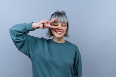 Genç bir kadın sade bir arka planın önünde duruyor. Gülümsüyor ve barış işareti olarak iki parmağını kaldırıyor. Saçı şekilli ve basit bir kazak giyiyor..