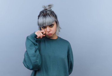 Yeşil bir kazak giymiş açık mavi bir duvarın önünde duran genç bir adam. Gümüş saçları var ve gözlerini parmaklarıyla dürterken hüzünlü bir ifade sergiliyorlar..