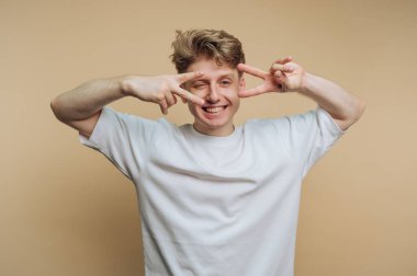 Genç bir adam gülümser ve kahverengi bir duvara karşı poz verirken elleriyle barış işaretleri yapar. Bu gündelik ortamda mutlu ve rahat görünüyor..