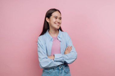 Kollarını kavuşturmuş genç bir kadın gülümsüyor. Açık mavi bir gömlek ve kot pantolon giyiyor. Arkaplan koyu pembe bir renktir. Bu sahne bir stüdyoda geçiyor..