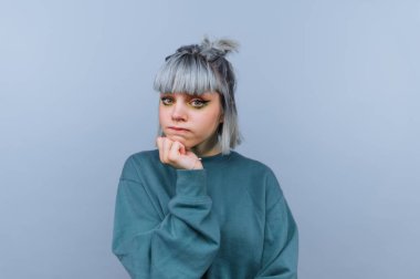 Gümüş saçlı genç bir kadın mavi bir duvarın önünde oturuyor. Yeşil bir kazak giyiyor ve eli çenesinde düşünceli bir yüz ifadesi sergiliyor..