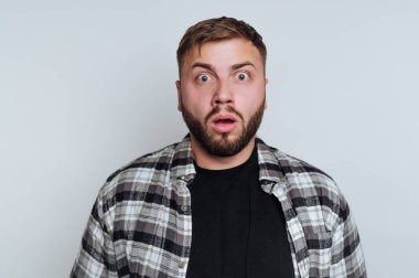 Bir adam şok olmuş bir ifadeyle içeride duruyor. Ağzı açık ve gözleri tamamen açık. Siyah bir tişörtün üzerine kareli bir gömlek giyer..