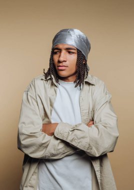 Örgülü saçlı genç bir adam kollarını kavuşturarak ayakta duruyor. Gümüş bir bandana ve basit bir gömlek giyiyorlar. Ayarlar sağlam bir renk arkaplanı.