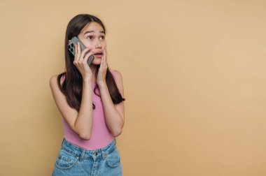 Bir kız akıllı telefonuyla konuşuyor. Yüzünde şok olmuş bir ifade var. Pembe gömlek ve kot pantolon giyerken sarı bir duvara dayanıyor..