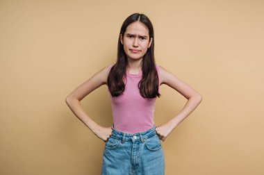 Genç bir kız elleri kalçasında ve endişeli bir ifadeyle ayakta duruyor. O, düz turuncu bir duvarın önündeki kameraya doğrudan bakar..