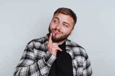 Düşünceli bir ifadeyle ayakta duran bir adam, parmakları çenesinde, siyah bir tişörtün üzerine ekose gömlek giymiş. Onun kısa bir sakalı var ve hafifçe yana bakıyor..