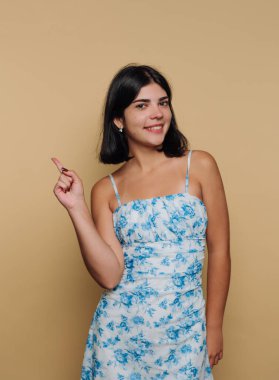 Genç bir kadın sağlam bir duvarın önünde duruyor. Çiçek desenli bir elbise giyiyor ve parmağıyla işaret ederken gülümsüyor. Ayar eğlenceli bir anı yansıtıyor..