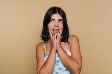 Elleri yüzünde ve neşeli bir bakışla genç bir kadın ayakta duruyor. Koyu saçlı ve mavi çiçekli bir bluz giyiyor. Arkaplanın düz bir rengi var.