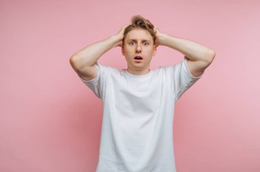 Elleri başının üstünde duran bir adam şaşırmış görünüyor. Arka plan pembe. Düz beyaz bir gömlek giyiyor. Dekor kapalı gibi görünüyor..