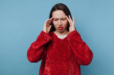 Uzun saçlı bir kadın açık mavi bir duvarın karşısında duruyor. Kırmızı bir kazak giyiyor ve kendini rahatsız hissediyor gibi görünüyor. İki eliyle başını tutuyor ve sıkıntı belirtileri gösteriyor..