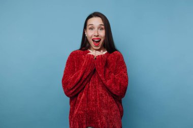 Elleri birbirine kenetlenmiş, gülümseyerek duran bir kadın. Kırmızı bir kazak giyiyor ve uzun saçları var. Arka plan mavi. Neşeli ve heyecanlı görünüyor..