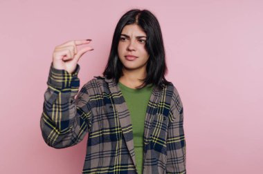 Bir kız pembe bir duvarın önünde duruyor, parmaklarını birbirine yaklaştırarak küçük bir bedeni gösteriyor. Yeşil bir gömlek ve ekose ceket giyiyor ve yüzünde anlamlı bir ifade var..