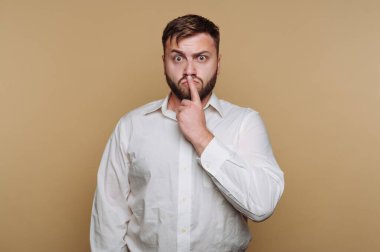 Bir adam sade bir arka planın önünde duruyor. Sakalı var ve beyaz bir gömlek giyiyor. Dudaklarında parmaklarıyla sessizliğini göstermek için bir jest yapıyor..