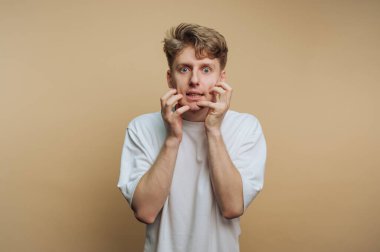 Yüzünde şaşkın bir ifadeyle bir adam ayakta duruyor. O korkunç bir ifade sergilerken elleri yanaklarını sıkıca tutuyor. Arkaplan basit bir bej rengi..