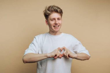 Stüdyoda hafif bir geçmişi olan genç bir insan duruyor. Gülümser ve ellerini kullanarak kalp şekli oluştururlar. Çekim sırasında neşe ve dostluğu yakalar..