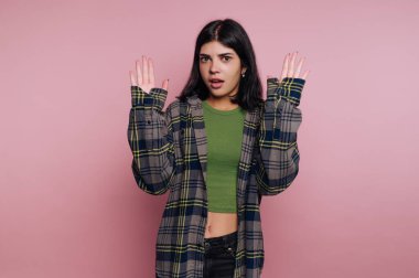 Elleri havada duran bir kadın, şaşırmış bir ifade sergiliyor. Damalı bir ceketin altına kesilmiş yeşil bir bluz giyiyor. Arka plan pembe ve içerisi de kapalı..