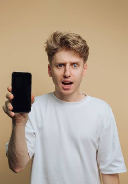 Genç bir adam düz bir arkaplanın önünde elinde şaşırmış bir ifadeyle akıllı bir telefonla duruyor. Saçı şekilli ve sade beyaz bir gömlek giyiyor..