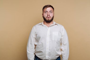Bir adam sabit duruyor, gözleri açık ve yüzünde şaşırmış bir ifade var. O beyaz bir gömlek ve kot pantolon giyiyor. Arka plan katı bir açık renk..