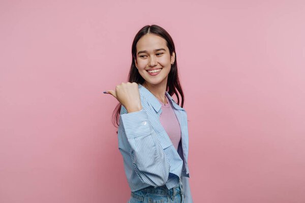 A young woman stands in front of a pink wall. She smiles and points to the side. Her casual outfit consists of a light shirt and a colorful shirt underneath. She looks happy.
