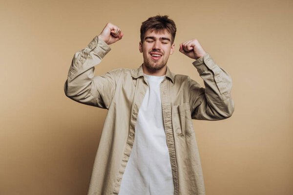A man raises his fists in celebration with a joyful expression. He wears a light-colored shirt over a t-shirt. The background is plain and neutral in tone.