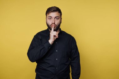 Siyah gömlekli adam sarı arkaplana karşı şşşş hareketi yapıyor..