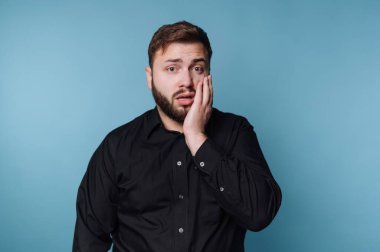 Siyah gömlekli bir adam şaşırmış görünüyor, yanağını tutuyor..