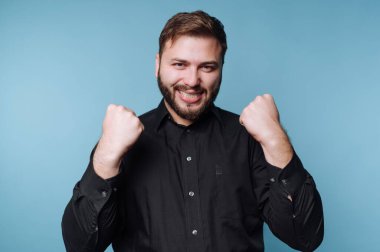 Siyah gömlekli bir adam gülümsüyor ve mavi arka planda yumruklarını kaldırıyor..