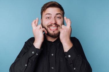 Siyah gömlekli bir adam mavi bir arka plana karşı umut dolu bir ifadeyle parmaklarını çaprazlıyor..