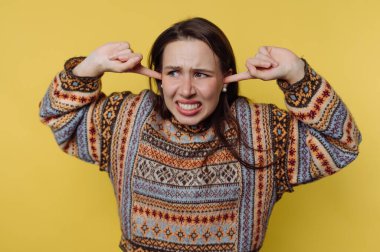 Sarı arka planda kulaklarını kapatan desenli kazaklı bir kadın.