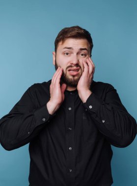 Siyah gömlekli bir adam endişeli görünüyor, mavi arka planda yüzüne dokunuyor..