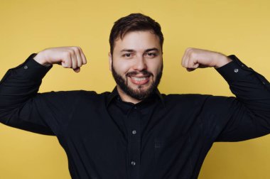 Siyah gömlekli gülümseyen adam sarı arka planda kaslarını esnetiyor..