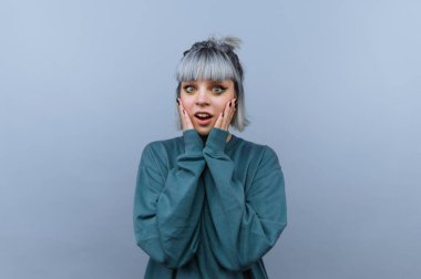 Gri saçlı, mavi arka planda deniz mavisi süveterli, şaşırmış genç bir kadın..