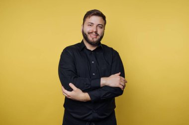 Sarı arka planda kolları çaprazlanmış, siyah gömlekli gülümseyen adam..