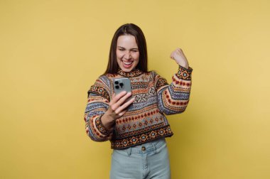 Sarı arka planda akıllı telefona bakarken kutlama yapan kadın.
