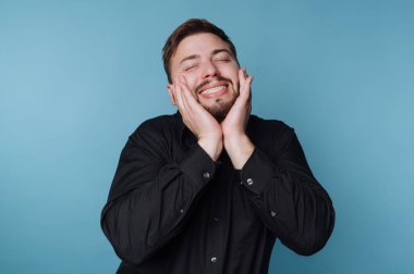 Elleri yanaklarında mavi arka planda gülümseyen siyah gömlekli bir adam..